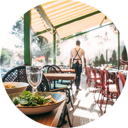 Slunná restaurace na terase s prostřenými stoly v popředí, na stole sklenice a talíř se salátem; v pozadí prochází číšník v zástěře venkovním prostorem.